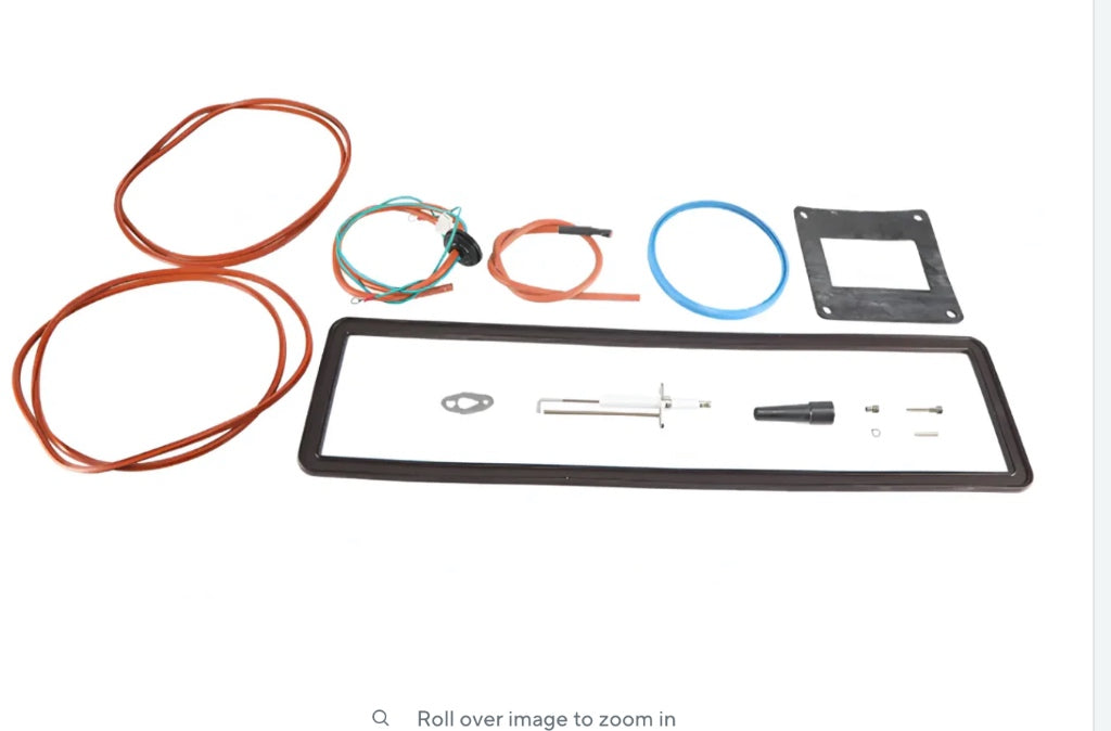 Studio product profile of a Patterson-Kelley 2600000344 spark igniter assembly, displaying its elongated white glazed ceramic insulation body, dual angled metal electrode firing rods, and a specialized integrated flat metal mounting bracket flange on a solid white background.